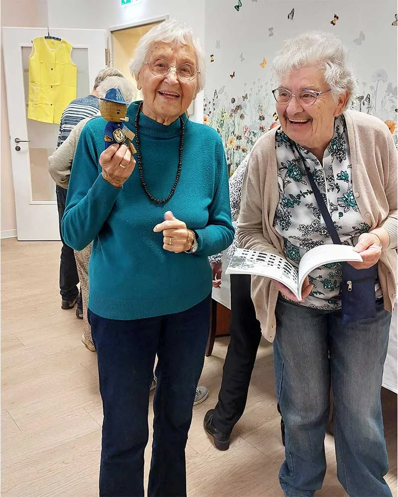 Zwei Damen stehen nebeneinander und lachen ausgelassen. Sie haben am Tag unserer DDR-Ausstellung in der Tagespflege Bücher und eine kleine Firgur mitgebracht, die sie an die Zeit von früher erinnern. 